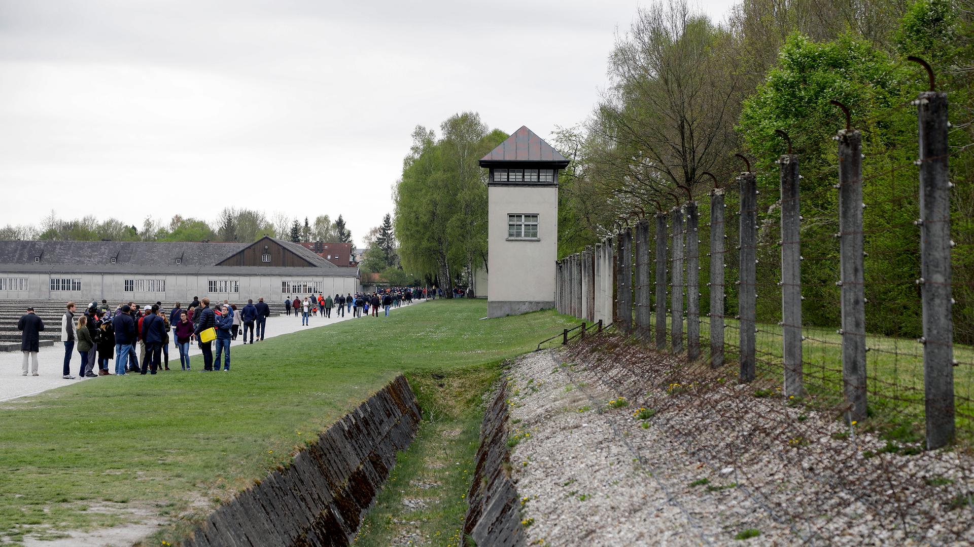 Blick auf den Wachturm des ehemaligen Konzentrationslagers Dachau mit Besuchern, die sich entlag des Gehweges verteilen.