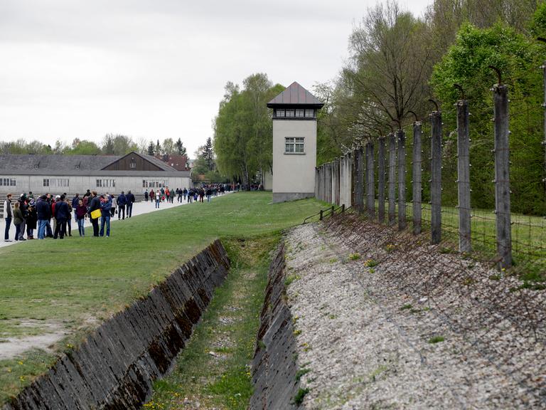 Blick auf den Wachturm des ehemaligen Konzentrationslagers Dachau mit Besuchern, die sich entlag des Gehweges verteilen.