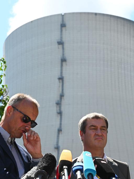 Markus Söder und Friedrich Merz stehen hinter Mikrofonen. Hinter ihnen der Kühlturm des Kernkraftwerks Isar 2.