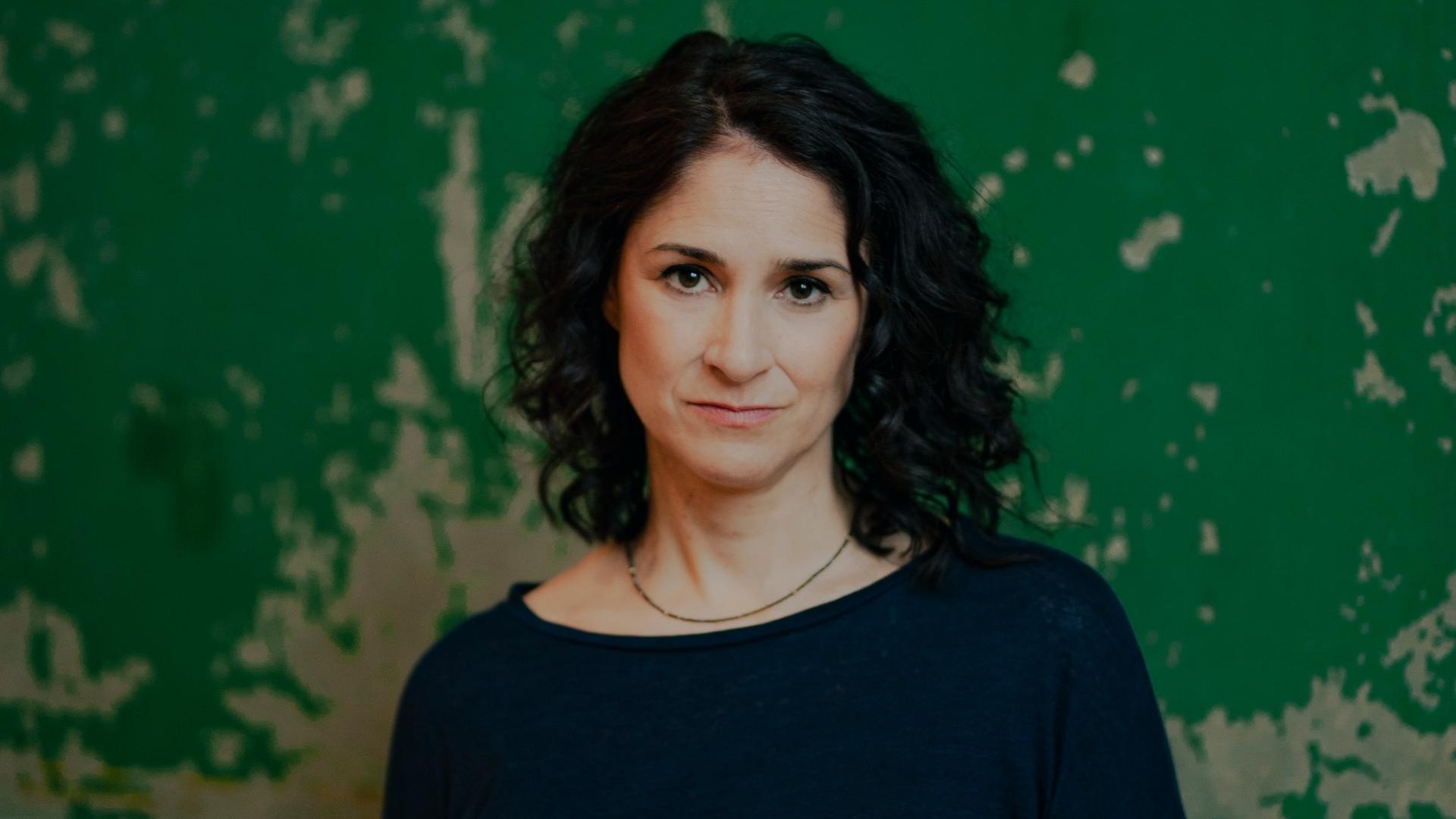 Porträtfoto der deutsch-ägyptischen Journalistin Annabel Wahba. Sie hat dunkles, langes Haar, trägt einen dunklen Pullover und steht vor einer grüngestrichenen Wand, von der die Farbe abblättert. Porträtfoto der deutsch-ägyptischen Journalistin Annabel Wahba. Sie hat dunkles, langes Haar, trägt einen dunklen Pullover und steht vor einer grüngestrichenen Wand, von der die Farbe abblättert.