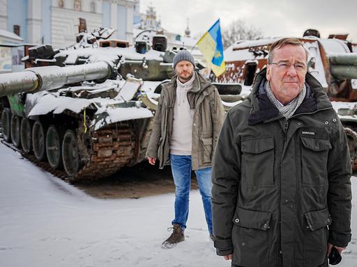 Boris Pistorius steht in einer olivgrünen Jacke vor zerstörten russischen Panzern in Kiew. Eine blau-gelbe Fahne ist im Hintergrund zu sehen.