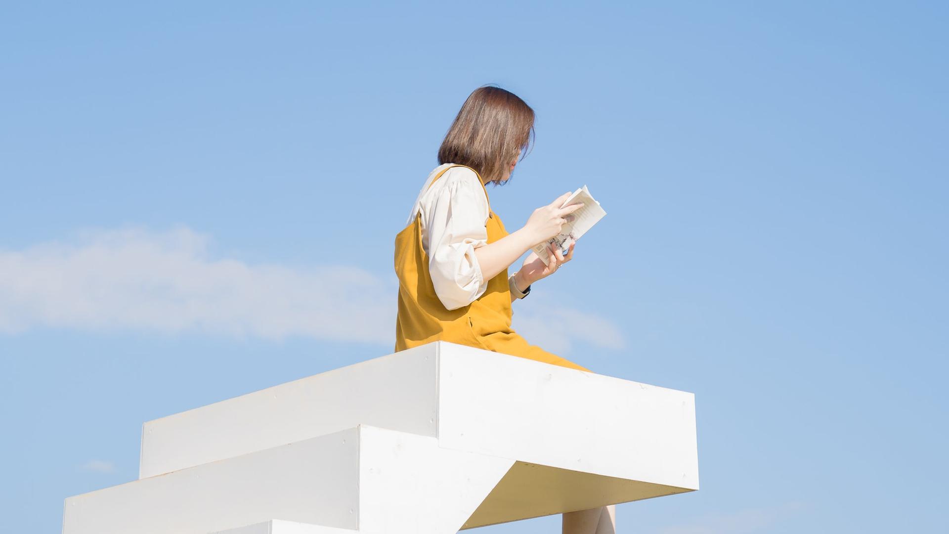 Eine weiße Treppe endet im Nichts vor blauem Himmel. Auf der letzten Stufe sitzt eine Person mit baumelnden Beinen und liest ein Buch. Eine weiße Treppe endet im Nichts vor blauem Himmel. Auf der letzten Stufe sitzt eine Person mit baumelnden Beinen und liest ein Buch.