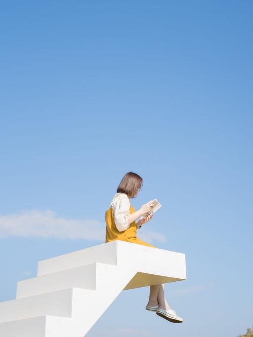 Eine weiße Treppe endet im Nichts vor blauem Himmel. Auf der letzten Stufe sitzt eine Person mit baumelnden Beinen und liest ein Buch.