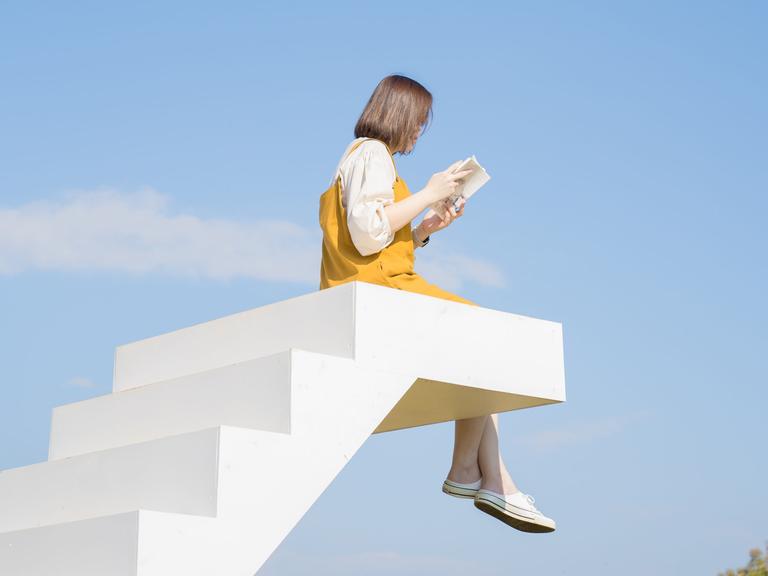 Eine weiße Treppe endet im Nichts vor blauem Himmel. Auf der letzten Stufe sitzt eine Person mit baumelnden Beinen und liest ein Buch.