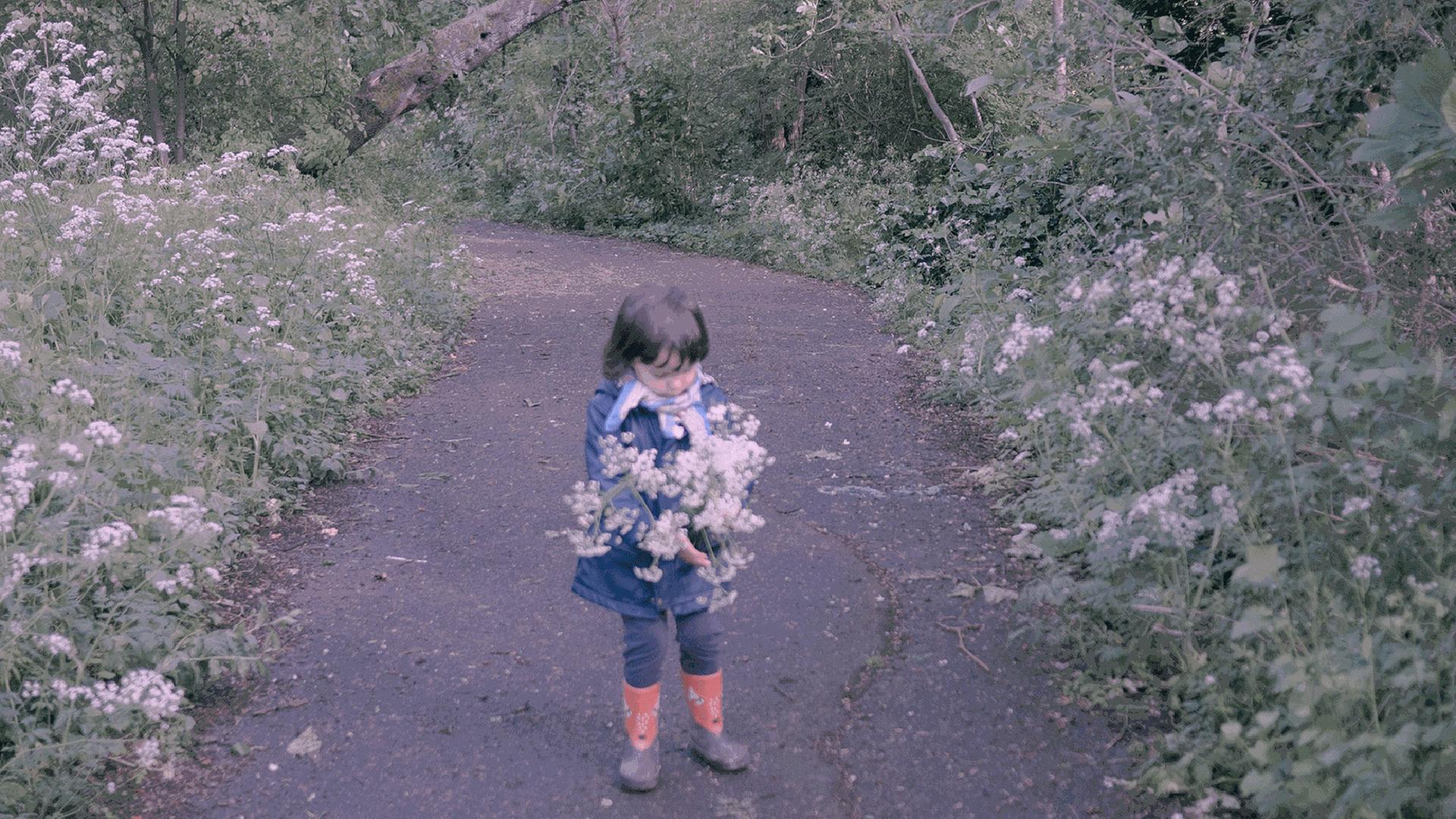 Zu sehen: Auf einem Weg in einem Park steht ein kleines Mädchen mit Blumen. Zu sehen: Auf einem Weg in einem Park steht ein kleines Mädchen mit Blumen.