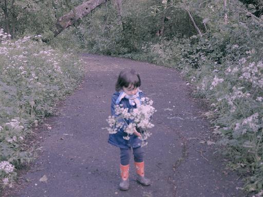 Zu sehen: Auf einem Weg in einem Park steht ein kleines Mädchen mit Blumen. 