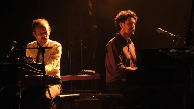 Zwei in sepiafarbenes Licht gehüllte Pianisten sitzen hinter ihren Instrumenten auf einer Bühne mit dunklem Hintergrund.