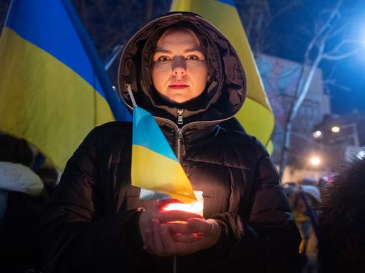 Zum Jahrestag des russischen Angriffs auf die Ukraine hält eine junge Frau bei einer Kundgebung in Montreal die ukranische Flagge und eine Kerze in der Hand, sie blickt in die Kamera. Im Hintergrund sind große ukrainische, gelb-blaue Flaggen zu sehen.