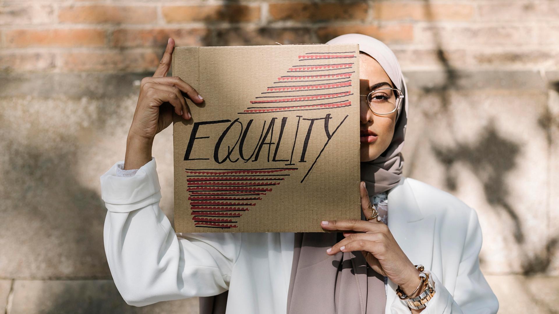 Eine junge Frau mit Kopftuch hält ein Schild vor ihr Gesicht, auf dem „Equality“ steht.