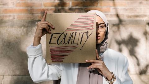 Eine junge Frau mit Kopftuch hält ein Schild vor ihr Gesicht, auf dem „Equality“ steht.
