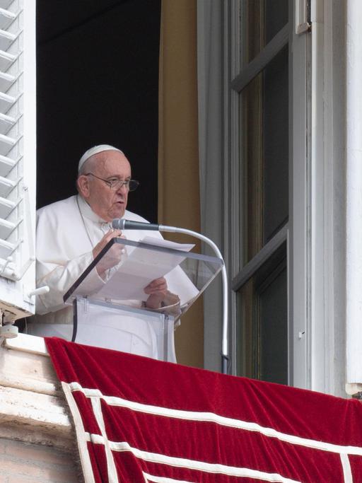 Papst Franziskus steht in weißem Gewand an einem durchsichtigen Rednerpult mit Mikrofon an einem offenen Fenster und spricht mit mehreren Seiten Papier in den Händen.
