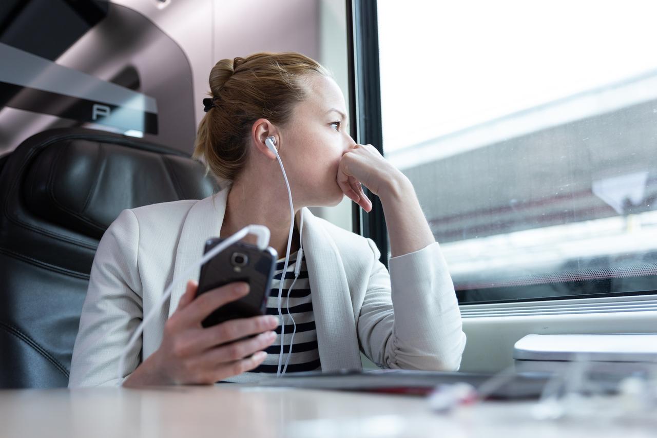 Eine Frau sitzt in der Bahn am Fenster mit Kopfhörern in den Ohren, die mit ihrem Handy verbunden sind. Eine Frau sitzt in der Bahn am Fenster mit Kopfhörern in den Ohren, die mit ihrem Handy verbunden sind.