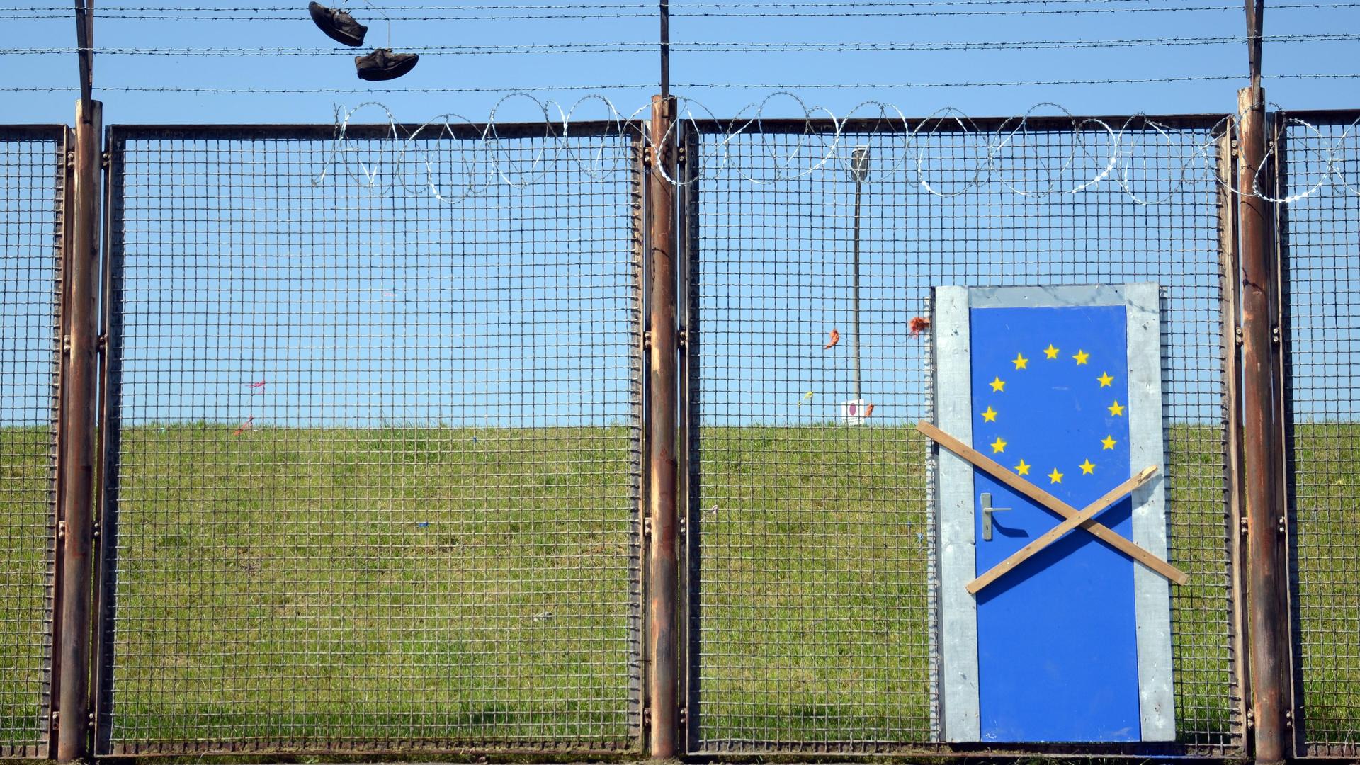 Symbolbild EU-Außengrenze: Zaun mit einer blauen EU-Tür, die mit zwei Holzbalken verbarrikadiert ist