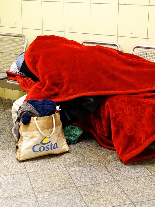Obdachloser zugedeckt mit einer roten Decke schläft auf einer Bank in der U-Bahnstation Leopoldplatz. 