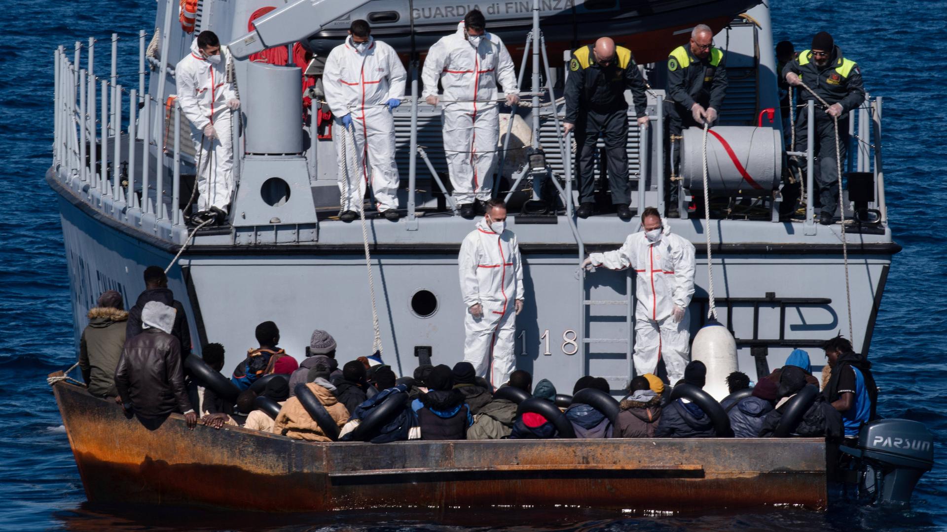 Helfer in weißen Schutzanzügen an Bord eines Bootes der italiensichen Küstenwache kümmern sich um Menschen, die in einem rostigen Boot auf See sitzen.