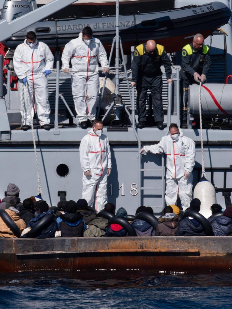 Helfer in weißen Schutzanzügen an Bord eines Bootes der italiensichen Küstenwache kümmern sich um Menschen, die in einem rostigen Boot auf See sitzen.