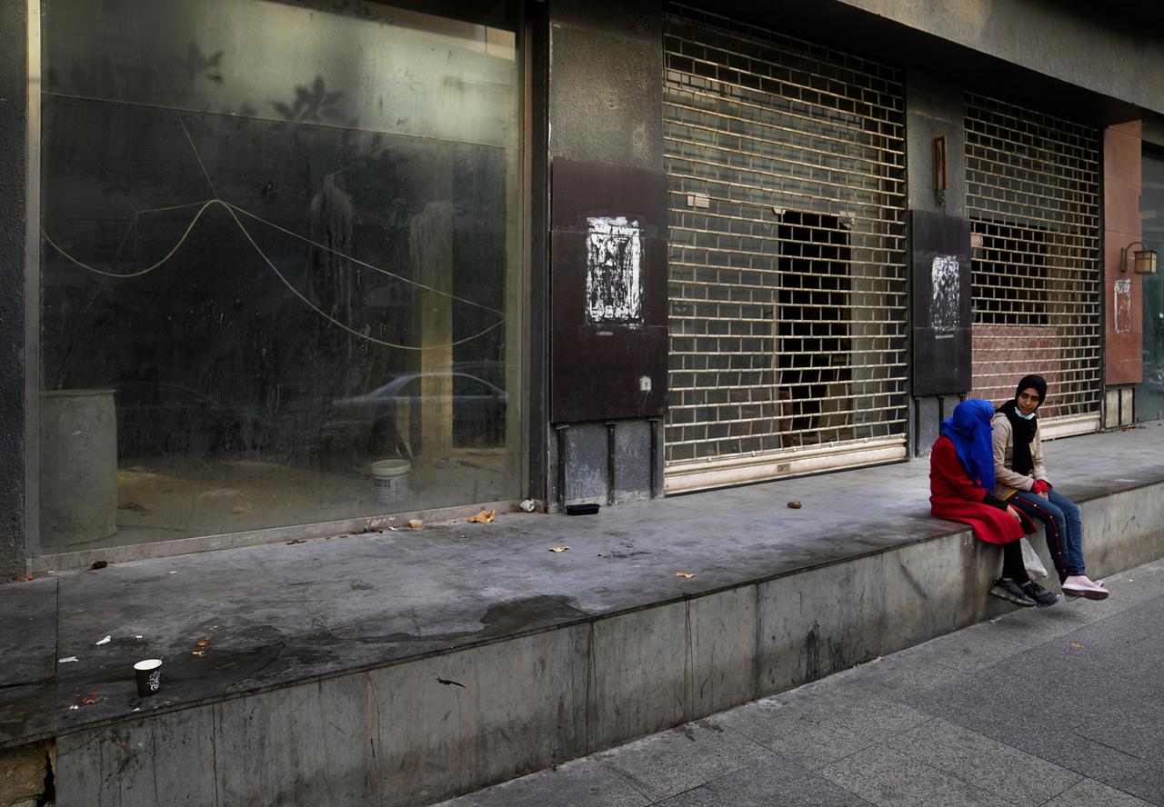 Zwei junge Mädchen mit Kopftuch sintzen inmitten einer verlassenen und schmutzigen Straße mit  geschlossenen Geschäften.