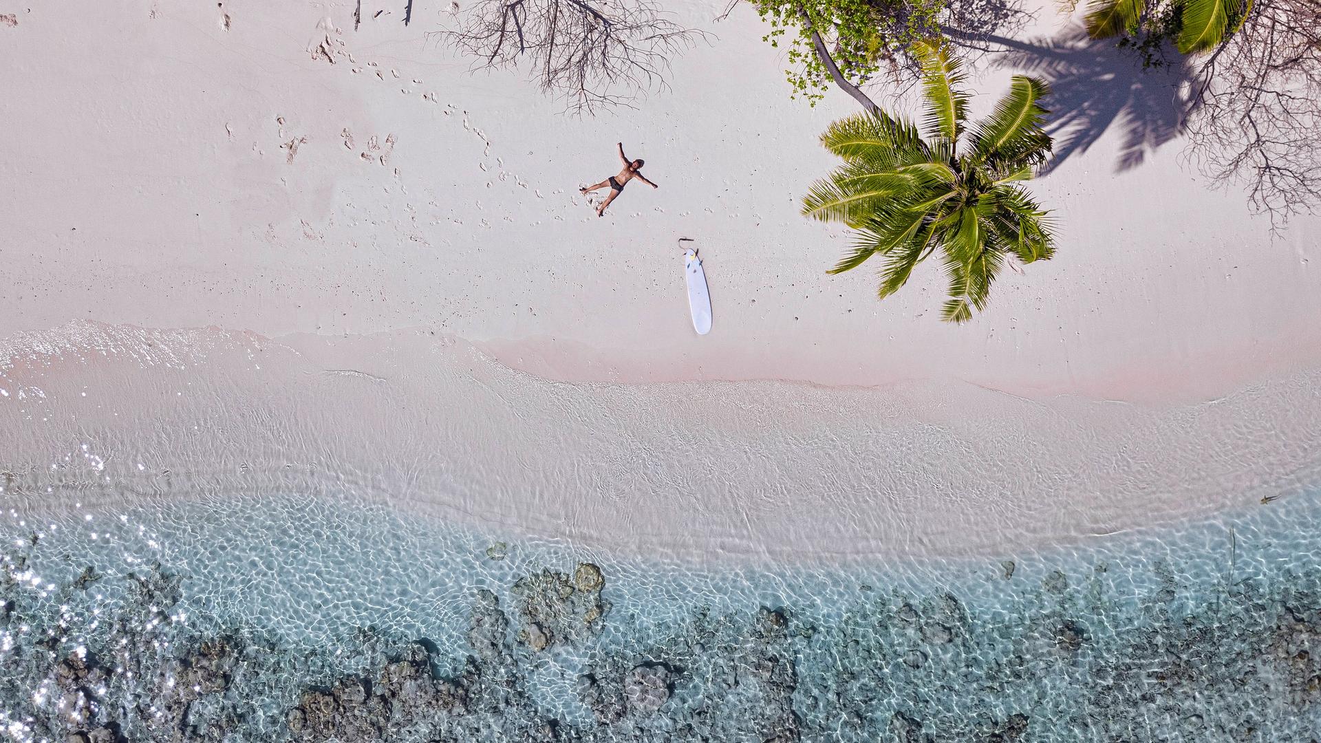 Ein Mann liegt mit einem Surfboard an einem einsamen Strand.