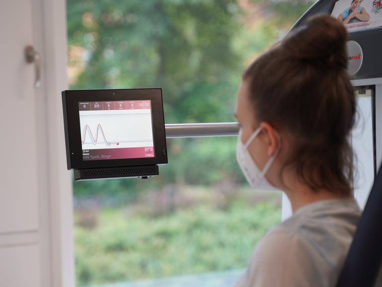 Die "Long Covid"-Patientin Birgit S. bei der Medizinischen Trainingstherapie im Unfallkrankenhaus Berlin (ukb). Sie trägt eine Maske und blickt auf einen Bildschirm mit einem Kurvenverlauf.
