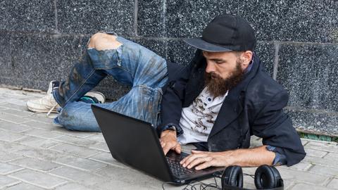 Ein junger Mann mit Baseballkappe liegt auf einem Bürgersteig auf dem Boden und bedient seinen Computer. 