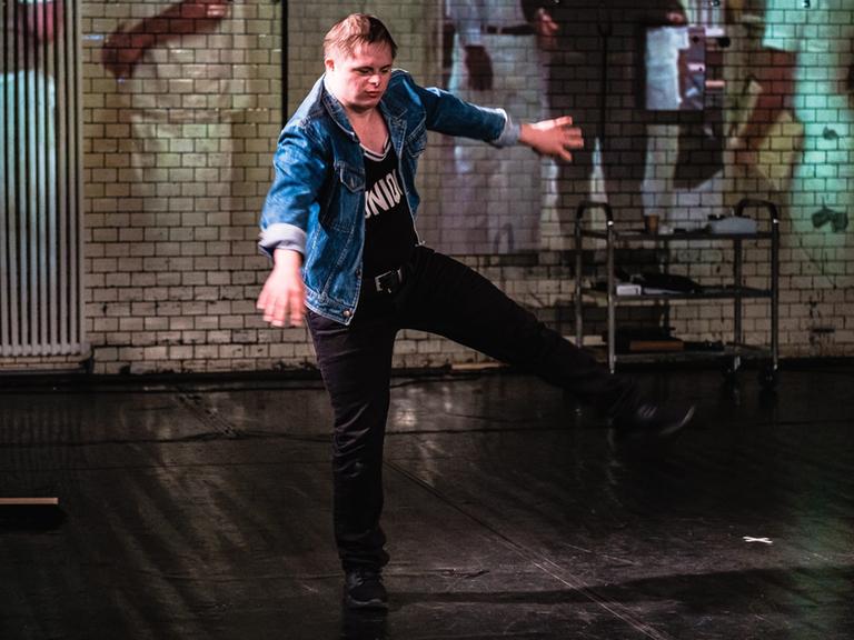 Ein Schauspieler in blauer Jeansjacke und dunklem T-Shirt balanciert auf einem Bein und rudert mit den Armen. Es ist ein Schauspieler der Theatergruppe RambaZamba in Berlin.