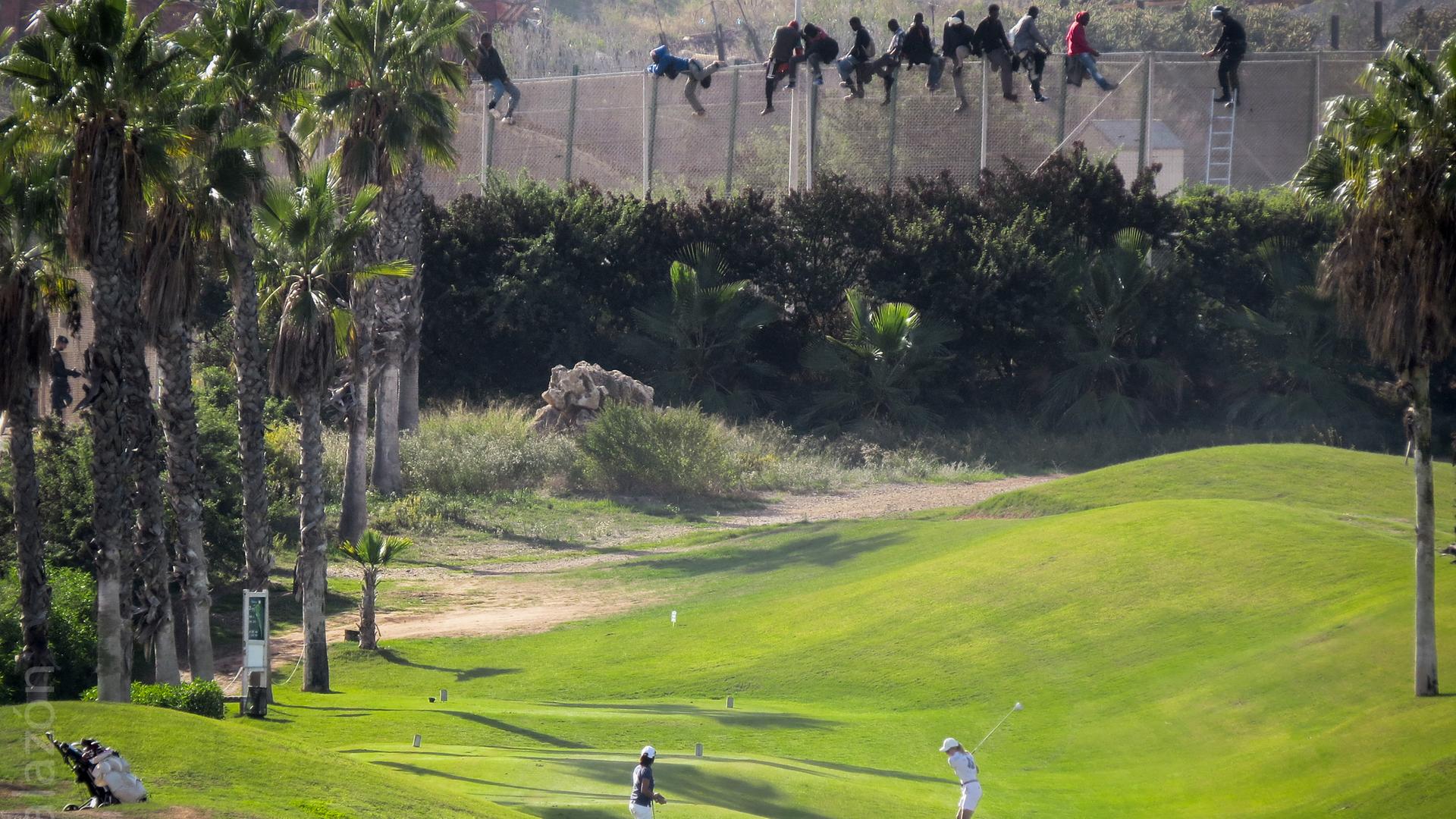 Ein Pärchen spielt unbekümmert Golf, obwohl direkt hinter ihm ein hoher Stacheldrahtzaun aufragt, ein Grenzzaun der spanischen Enklave Melilla in Marokko. Auf dem Zaun erkennt man einige afrikanische Männer, die gerade versuchen, hinüberzuflüchten. Ein Pärchen spielt unbekümmert Golf, obwohl direkt hinter ihm ein hoher Stacheldrahtzaun aufragt, ein Grenzzaun der spanischen Enklave Melilla in Marokko. Auf dem Zaun erkennt man einige afrikanische Männer, die gerade versuchen, hinüberzuflüchten.