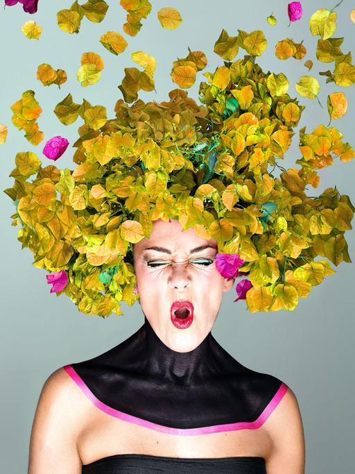 Eine Frau mit knallig geschminkten Mund scheint ein "O" zu singen, während ihr Kopf von gelblich-grünen und pinken Blättern umhüllt wird.