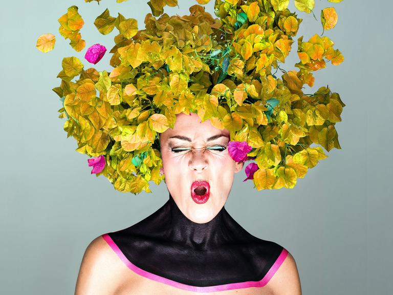 Eine Frau mit knallig geschminkten Mund scheint ein "O" zu singen, während ihr Kopf von gelblich-grünen und pinken Blättern umhüllt wird.
