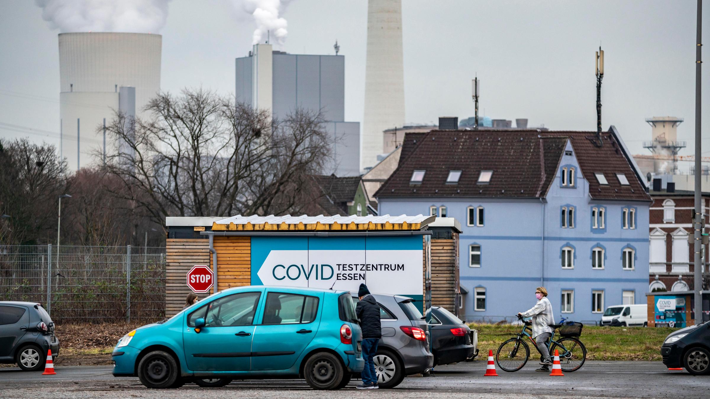 Corona-Drive-In-Testzentrum im Ruhrgebiet: Im Vordergrund sind Autos und ein Hinweisschild auf das Testzentrum zu sehen, im Hintergrund rauchen Kraftwerk-Schlote.