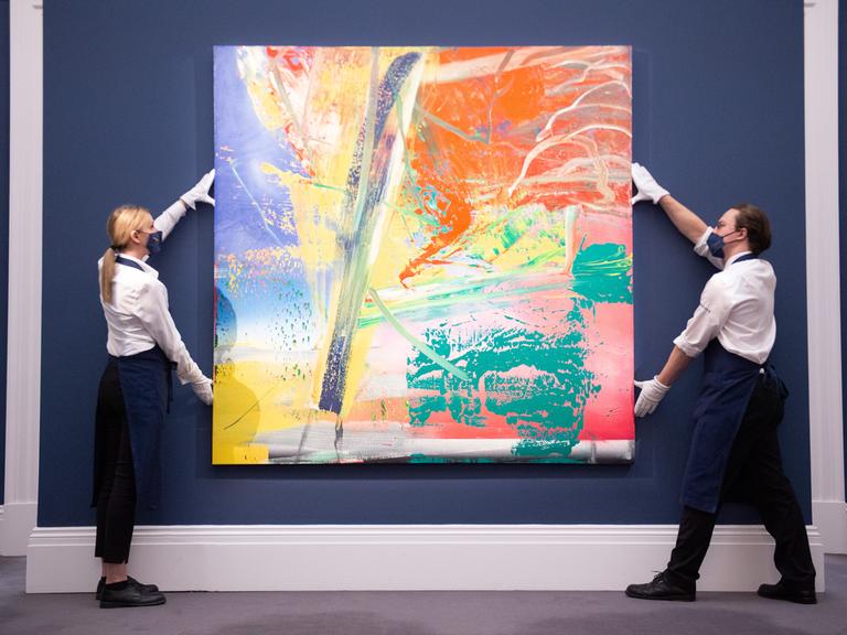 Zwei Personen halten das Geälde "S.D." von Gerhard Richter vor eine blaue Wand
