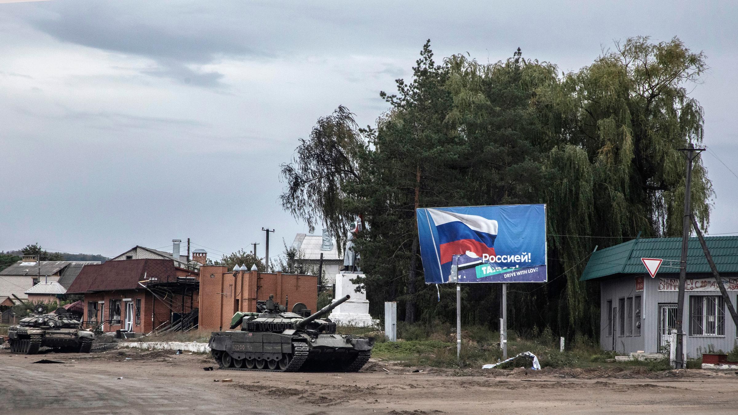 Ein zurück gelassener russischer Panzer auf einer zerstörten Straße in Izjum, Ukraine. Im Hintergrund ist ein Plakat mit der Flagge Russlands zu sehen.