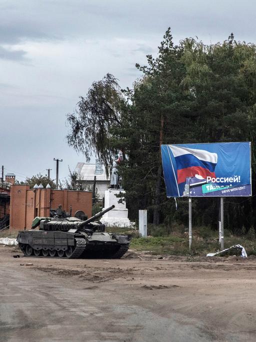 Ein zurück gelassener russischer Panzer auf einer zerstörten Straße in Izjum, Ukraine. Im Hintergrund  ist ein Plakat mit der Flagge Russlands zu sehen.