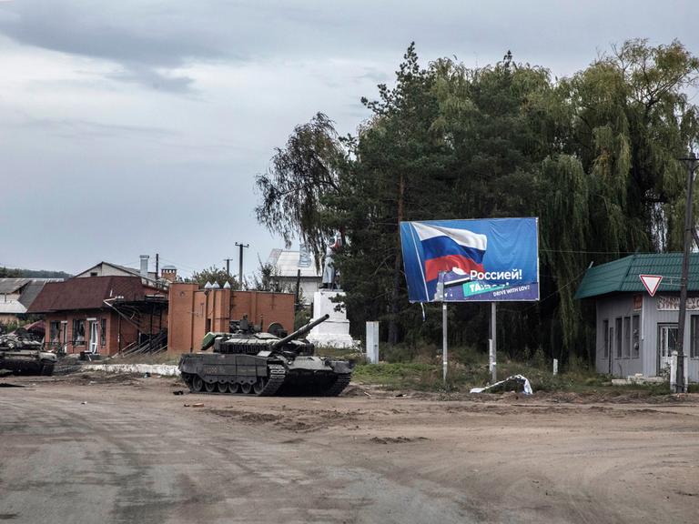 Ein zurück gelassener russischer Panzer auf einer zerstörten Straße in Izjum, Ukraine. Im Hintergrund  ist ein Plakat mit der Flagge Russlands zu sehen.