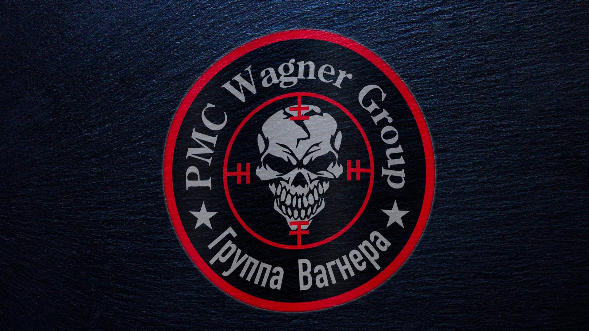 In einem inneren roten Kreis ist ein grinsender Totenkopf abgebildet, in einem äußeren die Schrift "PMC Wagner Group" sowie kyrillische Schrift. Der Hintergrund ist schwarz.