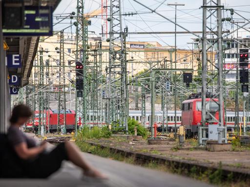 Eine Frau sitzt an einem Bahnsteig des Stuttgarter Hautpbahnhofs. Im Hintergrund sind Züge und Oberleitungen zu sehen. 
