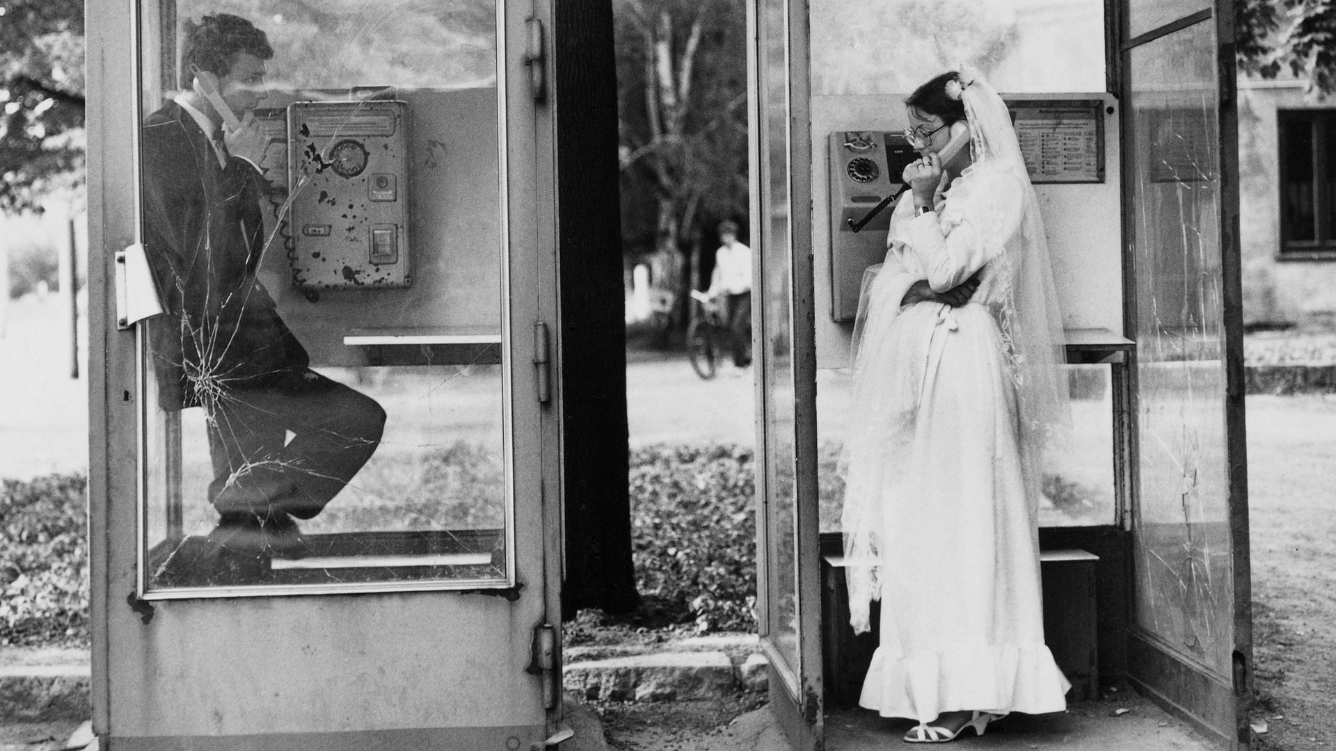 Ein Hochzeitspaar in zwei Telefonzellen in Leipzig, sie telefonieren nebeneinander miteinander, DDR, 1984. Ein Hochzeitspaar in zwei Telefonzellen in Leipzig, sie telefonieren nebeneinander miteinander, DDR, 1984.