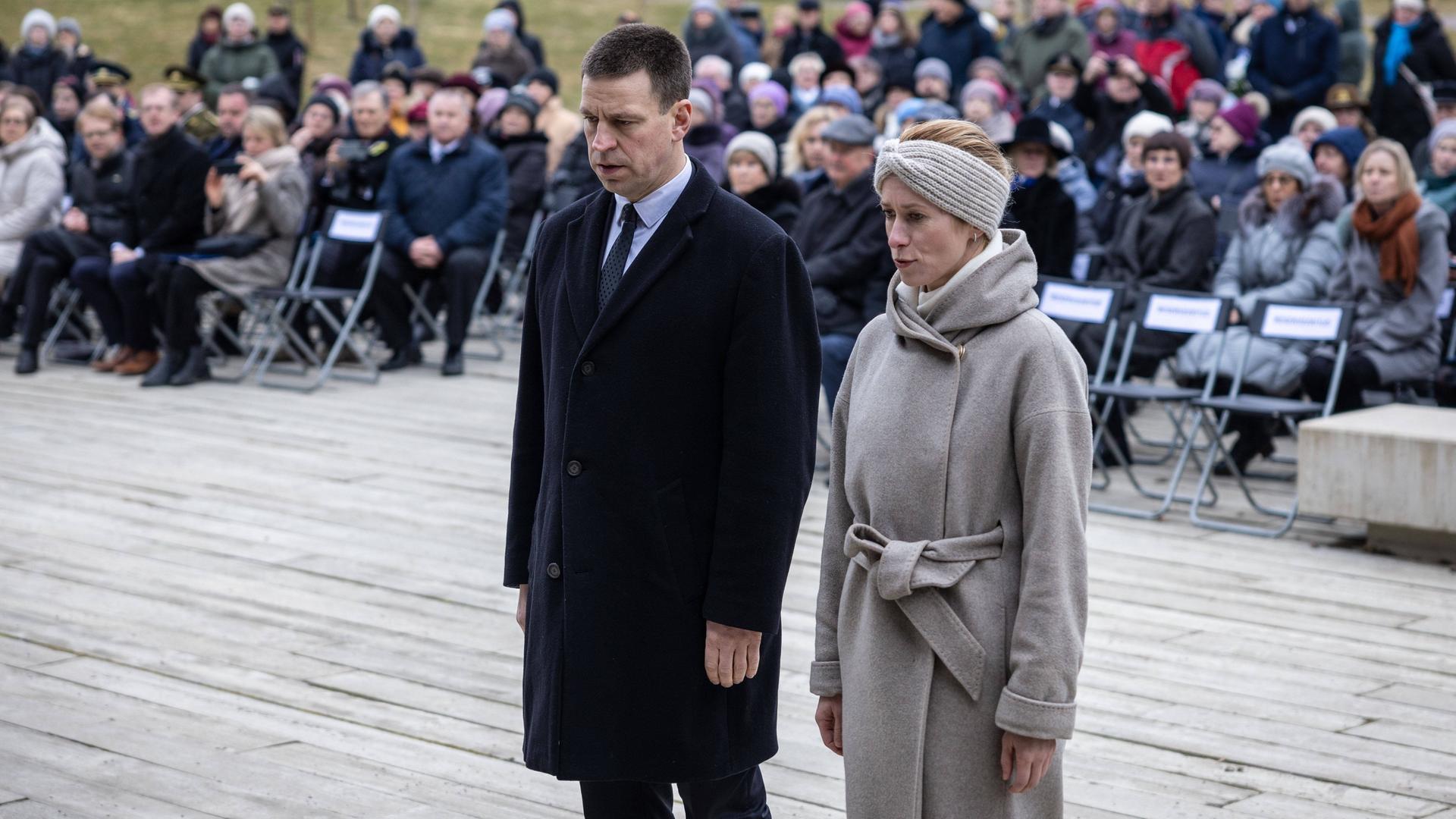 Die estische Ministerpräsidentin Kaja Kallas steht während einer Gedenkveranstaltung neben ihrem Amtsvorgänger Jüri Ratas. Beide blicken zu Boden.