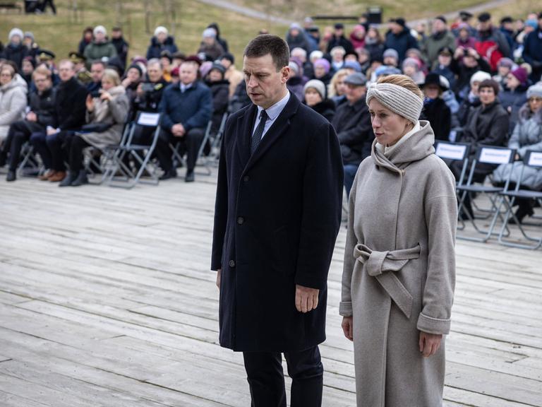 Die estische Ministerpräsidentin Kaja Kallas steht während einer Gedenkveranstaltung neben ihrem Amtsvorgänger Jüri Ratas. Beide blicken zu Boden.