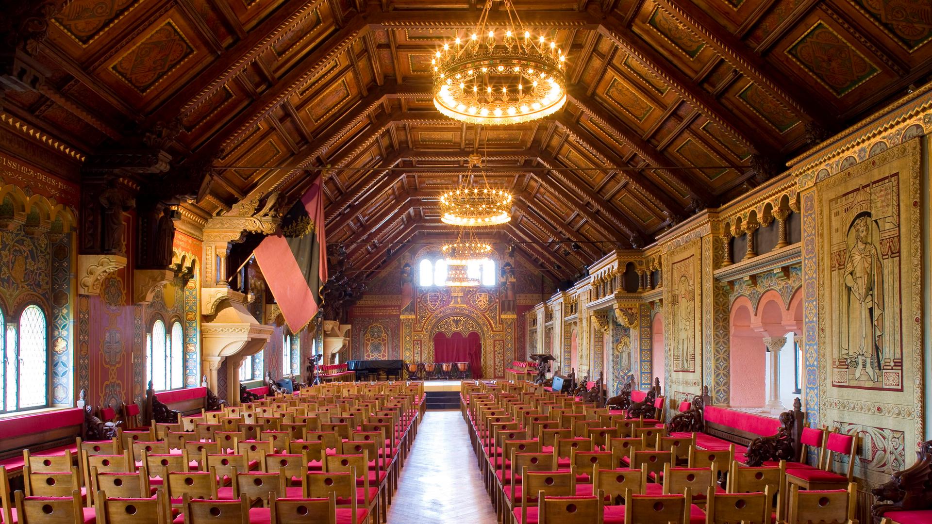 Blick in einen großen, historischen Saal mit hoher, dekorierter Holzdecke, imposanten Leuchten und roten Stühlen. Blick in einen großen, historischen Saal mit hoher, dekorierter Holzdecke, imposanten Leuchten und roten Stühlen.