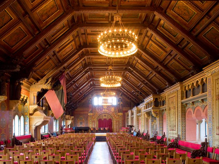 Blick in einen großen, historischen Saal mit hoher, dekorierter Holzdecke, imposanten Leuchten und roten Stühlen.