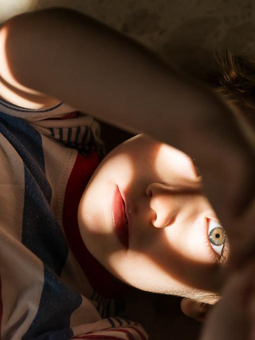 Ein Kind auf dem Rücken liegend von oben. Licht und Schatten auf seinem Gesicht, sodass der Eindruck entsteht, es liege in einer Höhle oder Grube.