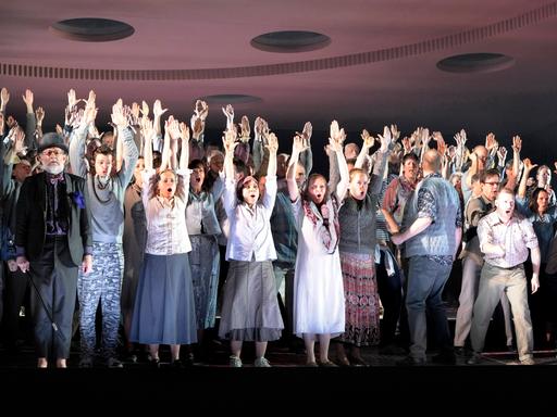 Etwa 50 Menschen stehen auf einer Bühne, nah an der Rampe und reißen die Arme in die Höhe. Es ist der Chor der Komischen Oper Berlin.