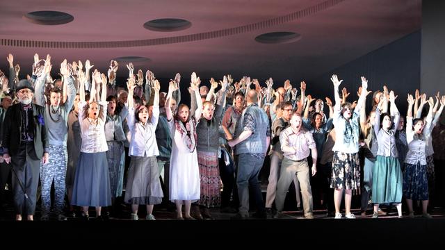 Etwa 50 Menschen stehen auf einer Bühne, nah an der Rampe und reißen die Arme in die Höhe. Es ist der Chor der Komischen Oper Berlin.