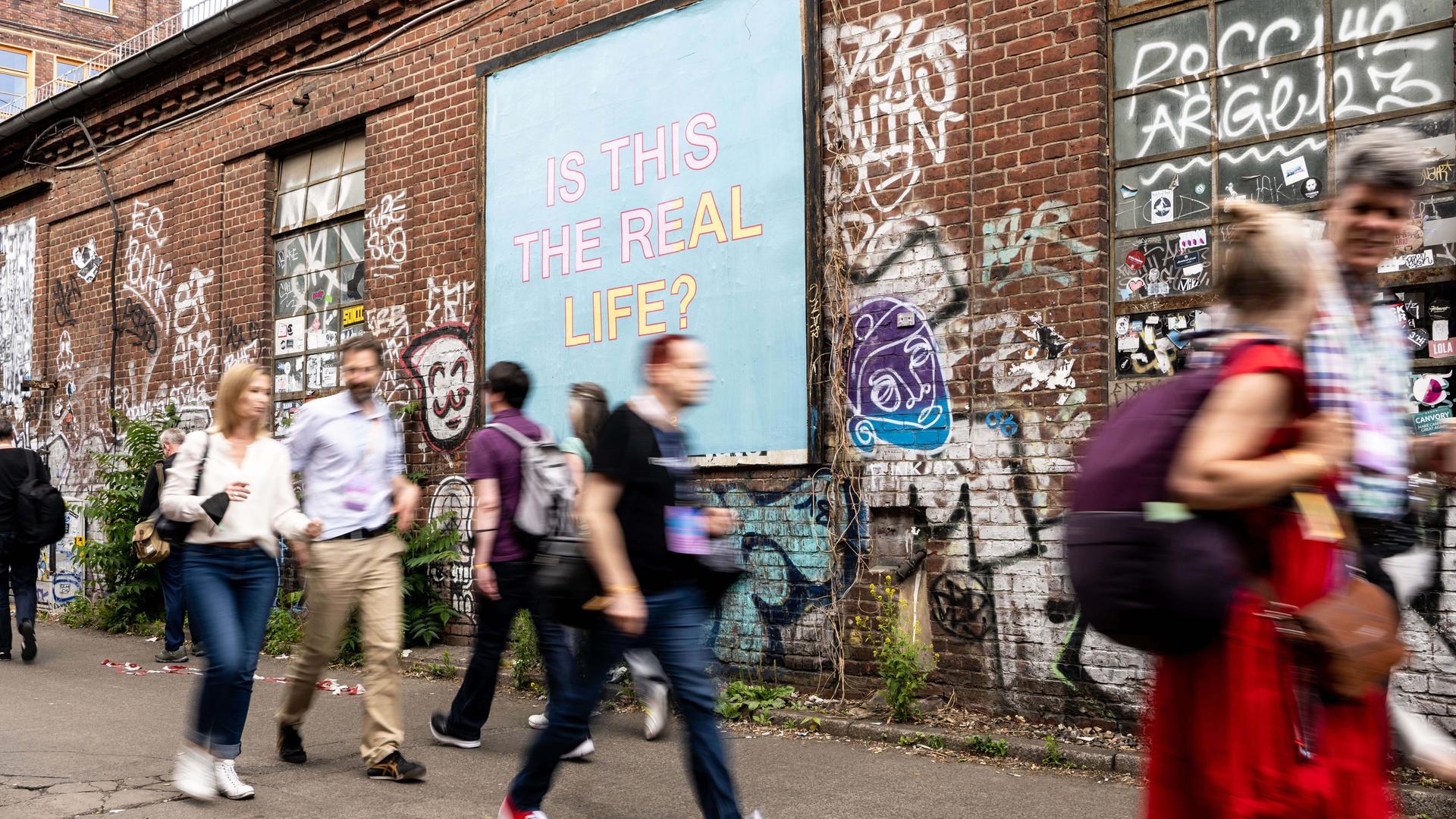 Verschwommene Menschen vor einem Plakat auf dem "Is This Real Life" steht. Verschwommene Menschen vor einem Plakat auf dem "Is This Real Life" steht.