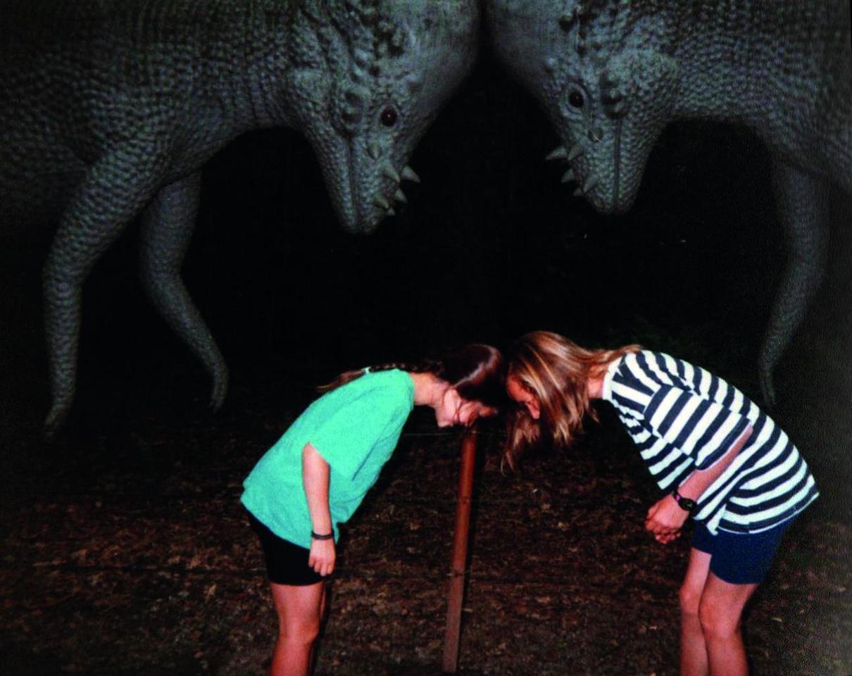 Zwei Mädchen imitieren die Pose der zwei Dinosauriermodelle hinter ihnen, die im Kampf ihre Köpfe gegeneinander drängen.