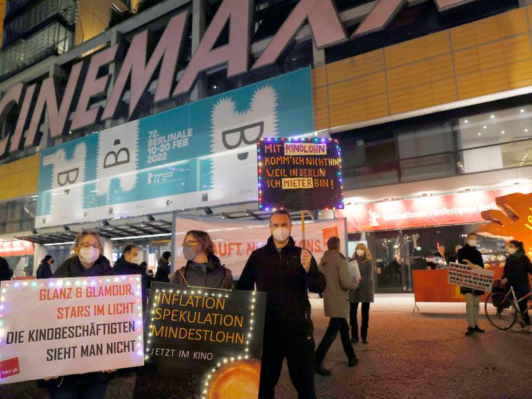 Mehrere Menschen stehen mit Plakaten vor einem Kino und demonstrieren. Auf einem steht beispielsweise: "Glanz und Glamour, Stars im Licht. Die Kinobeschäftigten sieht man nicht."