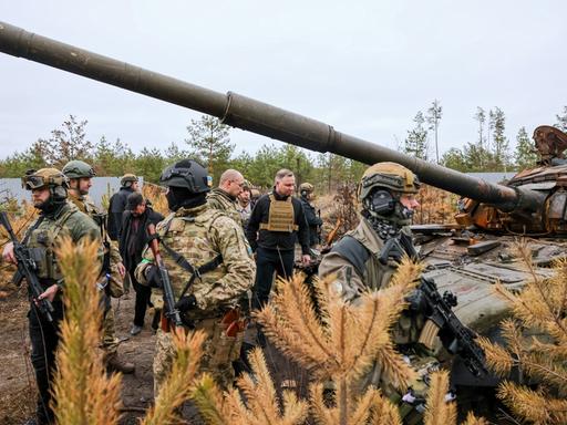 Ukraine: Andrzej Duda (M), Präsident von Polen, steht auf einem Feld neben einem zerstörten Panzer und ist dabei umgeben von bewaffneten Soldaten.