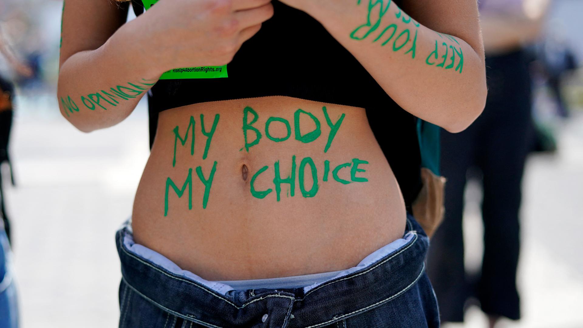 Eine Frau mit bauchfreiem Top nimmt an einer Demonstration teil. Auf ihrem Bauch sind mit grüner Farbe die Wörter "my body my choice" zu lesen.