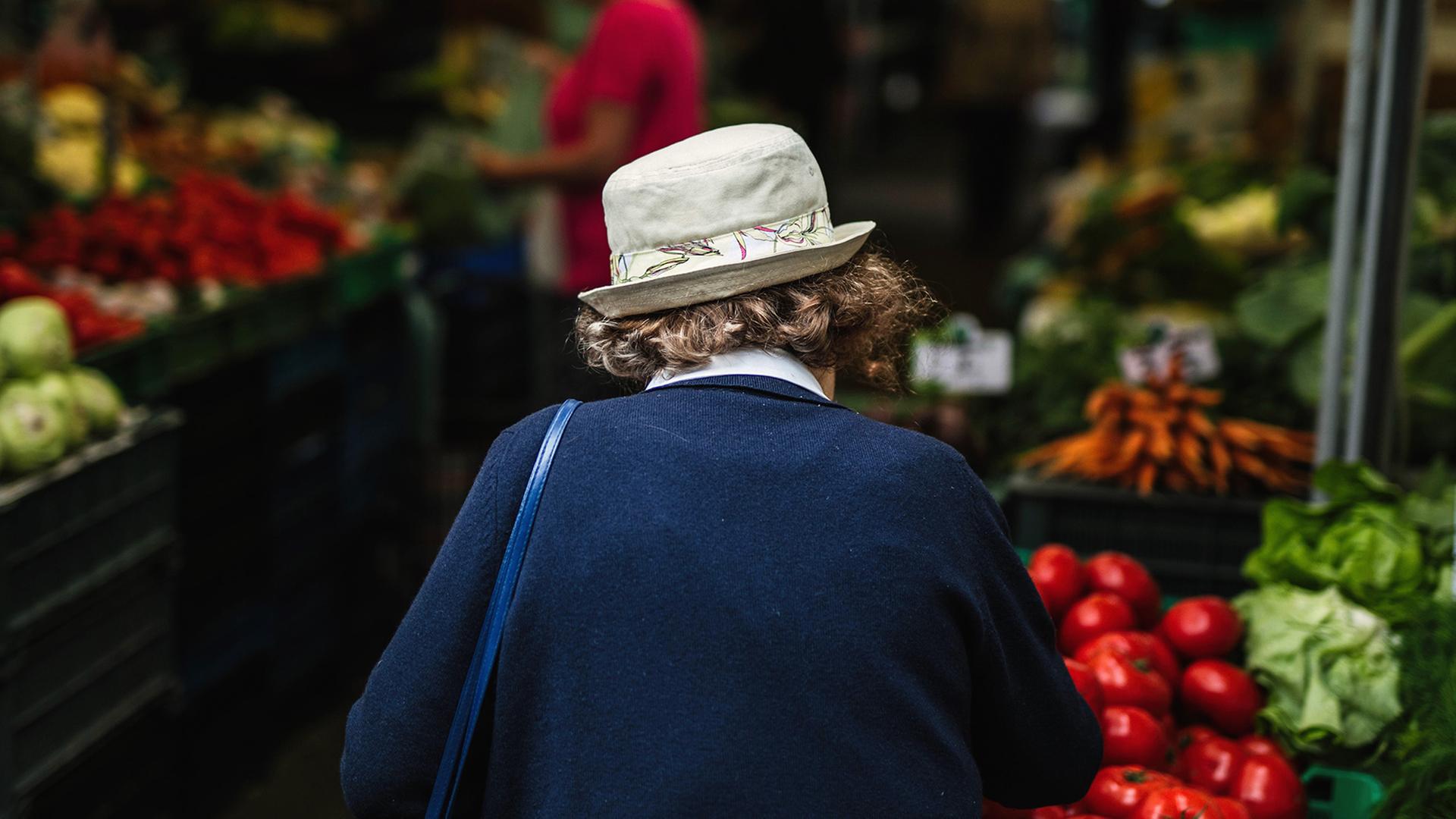 Eine ältere Frau mit Hut und dem Rücken zur Kamera geht über einen Gemüsemarkt. Eine ältere Frau mit Hut und dem Rücken zur Kamera geht über einen Gemüsemarkt.