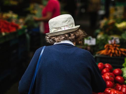 Eine ältere Frau mit Hut und dem Rücken zur Kamera geht über einen Gemüsemarkt. 